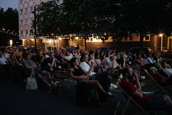 Foto: Viele Menschen sitzen in Stuhlreihen auf dem Vorplatz der Berlinischen Galerie und blicken nach vorne auf eine Leinwand, die nicht auf dem Bild zu sehen ist. Es dämmert langsam.